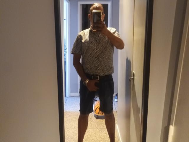 DanLemiaou, chauve, portant une chemise à carreaux, un short bleu, tenant un téléphone dans un miroir, couvrant ses fesses avec sa main, debout dans un couloir, sol recouvert de moquette, sac orange en arrière-plan..