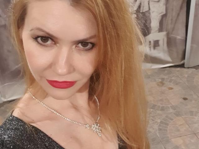 EvaNeon has red lipstick, sparkling eyes, and long blonde hair, wearing a glittery top with a necklace. Her tits are small, and she's looking directly at the camera.