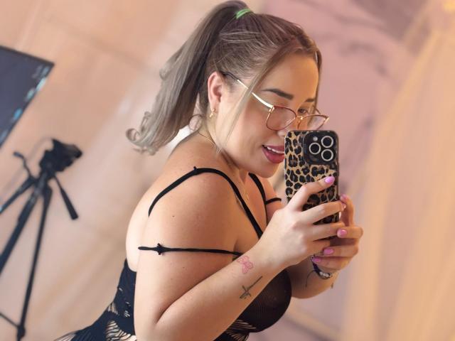 MiaBlondeHot, com óculos, rabo de cavalo, lingerie preta, segurando uma capa de celular com estampa de leopardo, esmalte rosa, tatuagens, sorrindo, seios médios, em pé, selfie, tripé ao fundo..