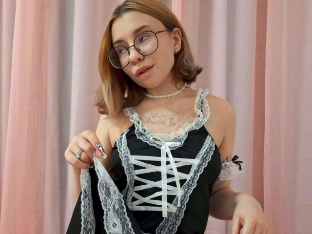 SugarNerdy, com óculos, cabelo loiro, vestido preto com cadarços, detalhes em renda branca, colar de pérolas, tatuagem no peito, segurando o vestido, cortinas rosa, fofa, sexy, seios pequenos, delicada, sorriso tímido..