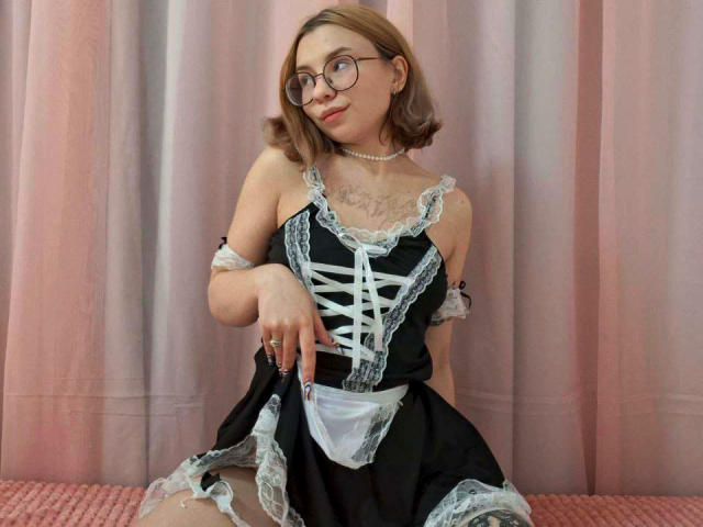 SugarNerdy, vêtue d'une tenue de femme de chambre noire et blanche en dentelle, assise avec un sourire mignon, des lunettes, des cheveux bruns courts, un collier et des tatouages sur la poitrine, montrant ses petites seins..