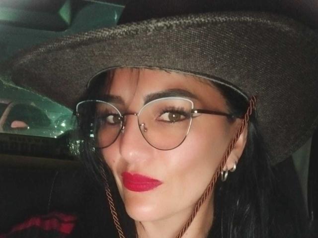 CrazyRousesex with dark hair, brown hat, red lipstick, and glasses, looking at camera, nose slightly upturned, earrings visible, black background, slightly shiny skin, confident expression.