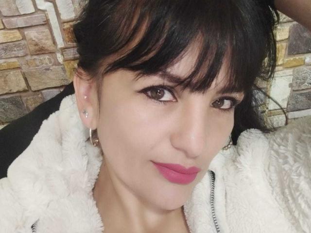 CrazyRousesex has black hair, brown eyes, pink lipstick, and a white fluffy jacket. She's wearing earrings and looking at the camera with a slight smile.