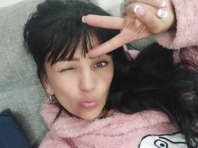 CrazyRousesex with black hair, one eye winking, pouting lips, wearing pink fuzzy sweater, making a peace sign with ringed finger, lying on gray couch.