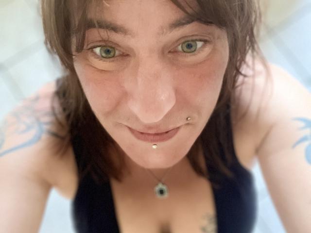 AcideCyprine with brown hair, blue eyes, nose piercings, and lip piercings, wearing a black top, showing small tattoos on her shoulders, and a necklace with a blue gem.