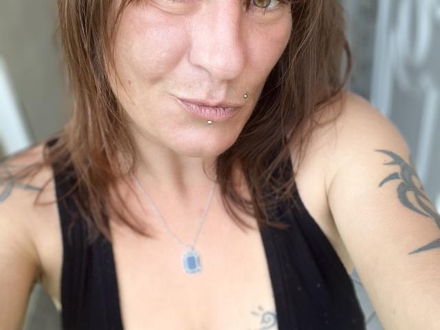 AcideCyprine has brown hair, piercings in her lower lip and nose, tattoos on her right arm, and wears a black sleeveless top, showing her small tits.