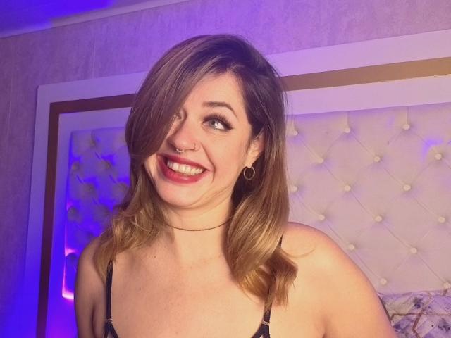 PaulettWatsoon with shoulder-length brown hair, smiling, nose ring, hoop earrings, red lipstick, black bra, choker, and cute face, in a dimly lit room with purple light.