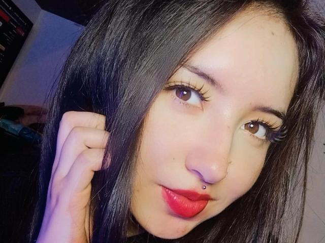 SusanMonroy has long black hair, brown eyes, and red lipstick with a small nose ring. She's touching her hair with her right hand, looking at the camera.