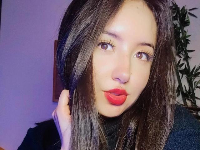 SusanMonroy has straight brown hair, brown eyes, red lipstick, and a nose ring. Her hand is near her head, looking at the camera with pouty lips.
