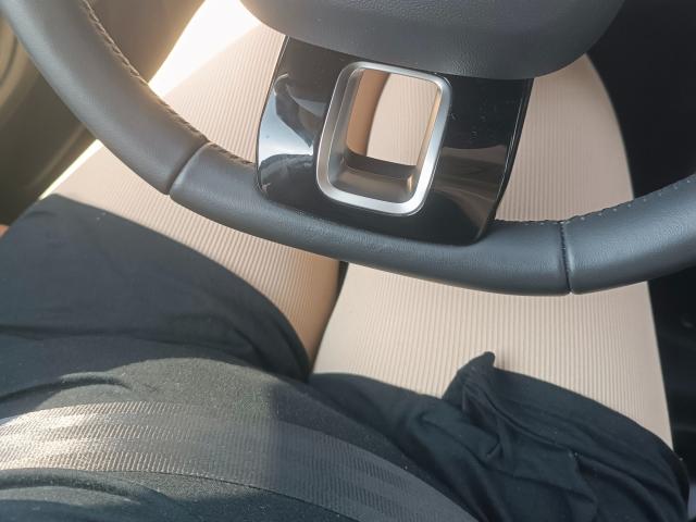 CurvyKompriGirl-hot's black leather steering wheel with a shiny metal logo, black pants, and beige seat, all shown in a tight, overhead shot.
