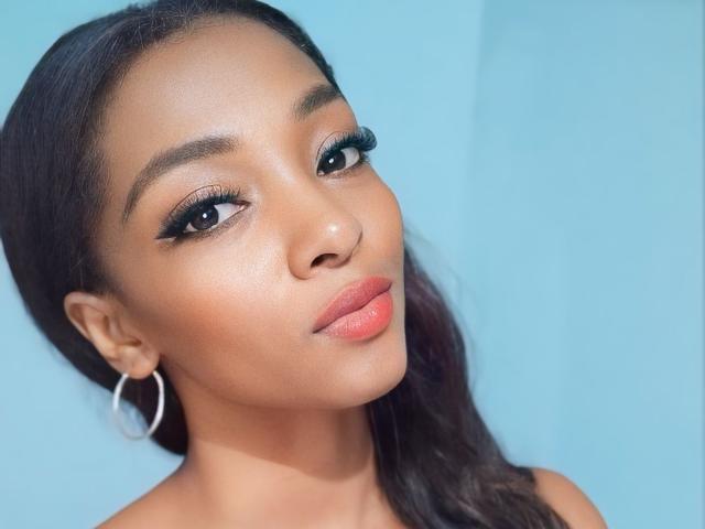 BrendaReyna has dark brown skin, full lips with pinkish-orange lipstick, thick black eyebrows, long eyelashes, and a side ponytail. She's wearing a silver hoop earring against a blue background.