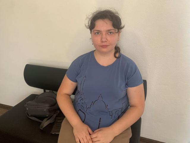 NeonilaLisova, a chubby woman with curly brown hair, blue line-drawing shirt, and tan pants, sits on a black chair with a gray backpack on a black table.