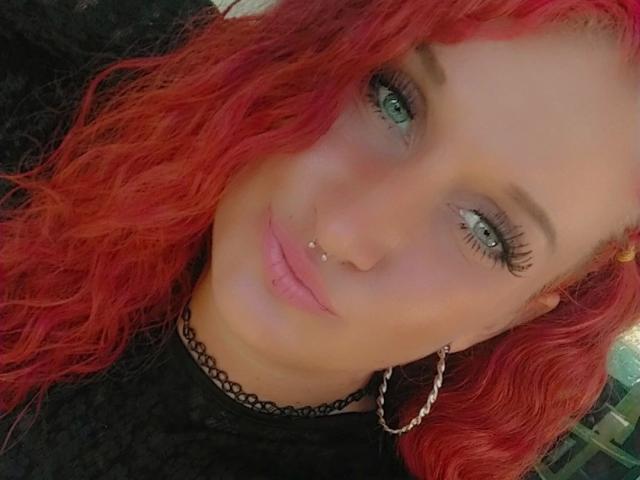 Licaink-hot has red curly hair, green eyes, black eyeliner, a nose piercing, a lip piercing, and wears a black choker with a silver chain earring.
