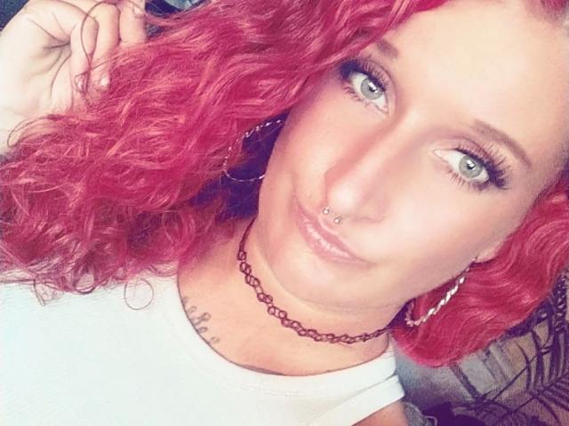 Licaink-hot with red curly hair, blue eyes, nose ring, and lip piercing, wearing a white shirt and choker, smiling at the camera. Boobs are not visible.