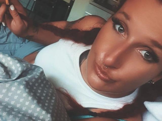 Licaink-hot with a nose piercing, wearing a white tank top, lying in bed, holding a cigarette, gray eyes, long red hair, tattoo on arm, and a pouty expression.