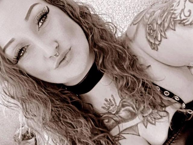 Licaink-hot with wavy hair, septum piercing, and tattooed arms, wearing a choker and lace-up top, showing her tits and a nipple piercing. Black-and-white photo.