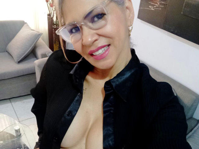 BrigiteWalsh with glasses, black button-up shirt, exposing her large boobs, smiling, pink lipstick, gold earrings, and light skin, in a cozy room with a gray sofa.