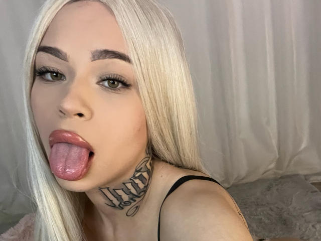 NickyBenz has blonde hair, green eyes, and a tattoo on her neck. She's sticking out her tongue with her pink lips and wearing a black strap.