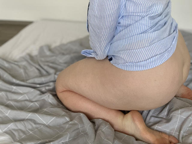 LilyyMoon's ass is exposed as she kneels on a bed, wearing an unbuttoned blue-striped shirt, with her legs folded under her. Her pale skin contrasts with the gray sheets.
