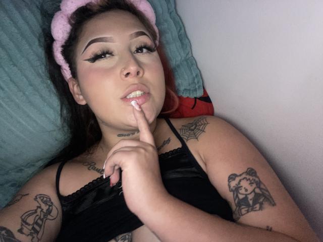 MorganLow lying on a bed, black top, pink headband, finger on lips, tattoos on arms, black eyeliner, nose ring, smiling, pink nail polish, red hair, white wall.