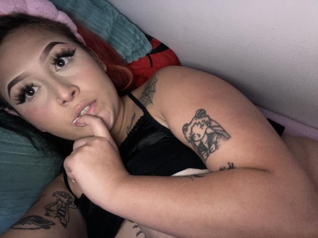 MorganLow, con tatuajes, está acostada en una cama, mordiéndose el dedo, vistiendo lencería negra, con pestañas largas y una expresión pensativa, mostrando sus pechos y tatuajes..