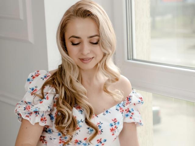 HottestSmile with long, wavy blonde hair, wearing a floral off-shoulder top, has a soft smile and closed eyes, standing near a window with natural light.