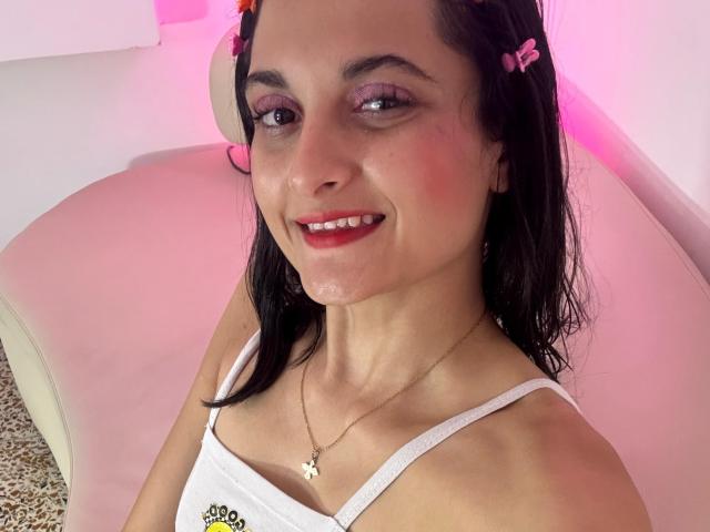 DulceSweety with black hair, pink butterfly hair clips, white tank top, red lipstick, pink eyeshadow, necklace, smiling, white couch, pink lighting, visible cum on her face.