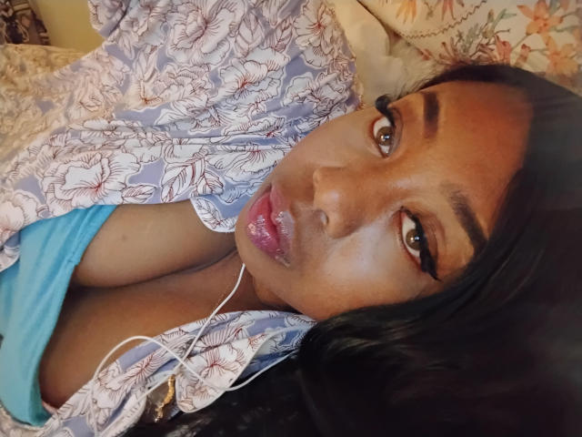 OliviaCambell with glossy brown eyes, full pink lips, and dark hair, lying down, wearing a floral shirt, blue top, and white earbuds, showing her large tits.