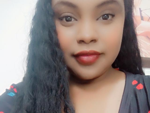 EbonyLatinAngel has smooth brown skin, long wavy black hair, and red lipstick. She's wearing a black shirt with red and blue accents, looking directly at the camera.