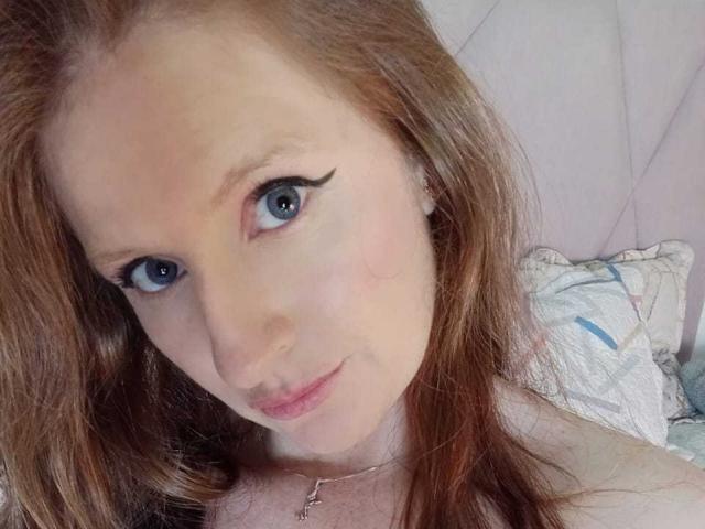 EnzumGinger has blue eyes, red hair, and a small nose, with subtle makeup including black eyeliner. She's wearing a necklace and lying on a pillow.