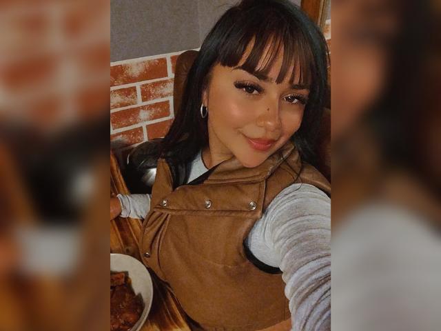 KarinaBlake with dark brown hair and bangs, smiling at the camera, wearing a brown leather vest over a white shirt, earrings visible, blurred figure in foreground.