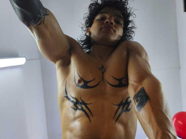 AlexBernal, shirtless, with curly black hair, tattoos on chest and arms, and a muscular, tan body, raising his right arm in a low-angle shot, showing small, dark nipples.