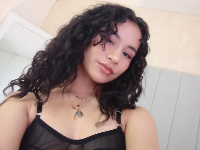 Heily Peterson, avec ses cheveux noirs bouclés, sa peau brun clair et son sourire discret, porte un haut noir transparent qui révèle ses petites seins, et elle porte un collier..