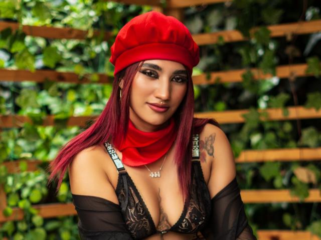 NemesisSinner with red beanie, red scarf, black lace bra, showing tits, red lipstick, tattoo on right shoulder, long red hair, black arm sleeves, and nose ring.