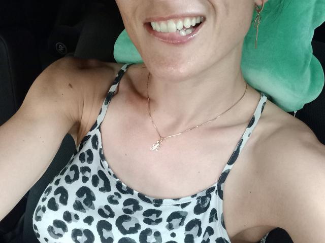 BellaJulls with a smiling face, showing teeth, wearing a leopard print tank top, and a silver necklace with a small "L" pendant, sitting in a car.