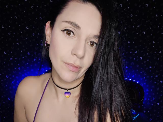 VelvetSkyX has pale skin, brown eyes, black hair, and wears a purple halter top with a black choker featuring a purple heart. She's looking directly at the camera.