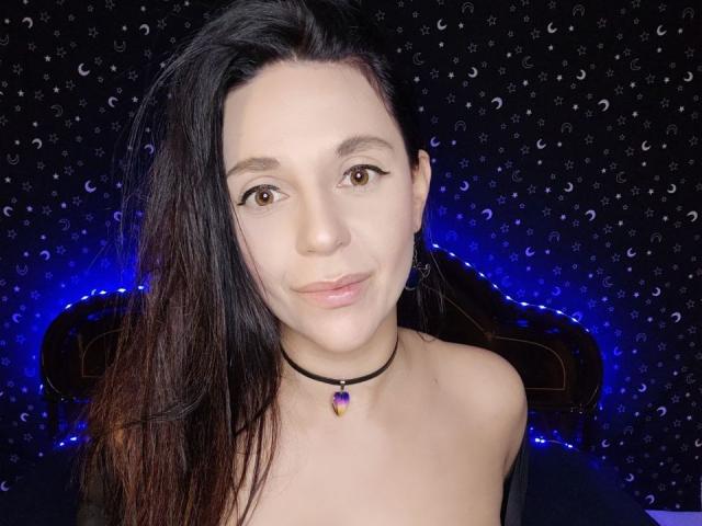 VelvetSkyX with long black hair, brown eyes, light skin, subtle smile, purple gem choker, bare shoulders, soft lighting, blue LED glow, patterned dark background.