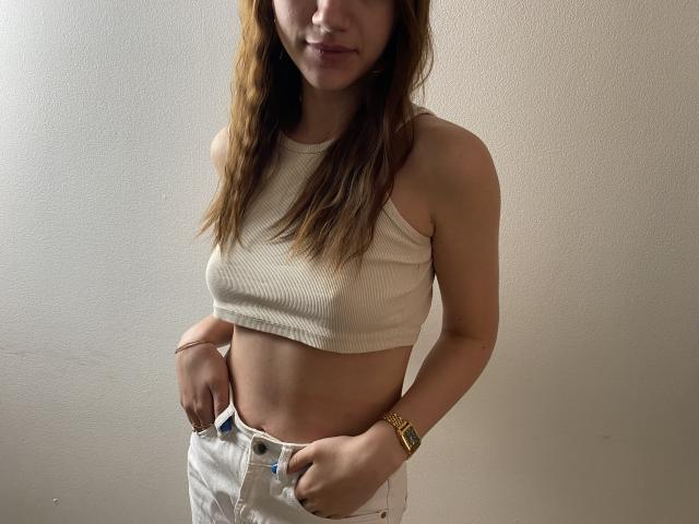 Twicksi with long brown hair, wearing a white crop top, showing her small tits, and white pants, hands in pockets, gold watch on left wrist, subtle smile, light skin.