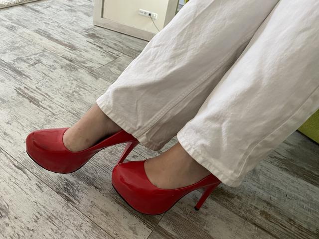 Twiksi's white pants and shiny red high heels with thick heels are on a wooden floor. Her pale skin is visible, showing just her feet and lower legs.
