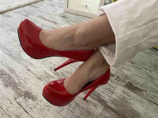 Los brillantes tacones rojos de Twiksi, con sus gruesos tacones de aguja, cruzaban sus piernas pálidas, que vestía con un vestido blanco, sobre un suelo de madera. Sus dedos estaban apuntando, mostrando los tacones..