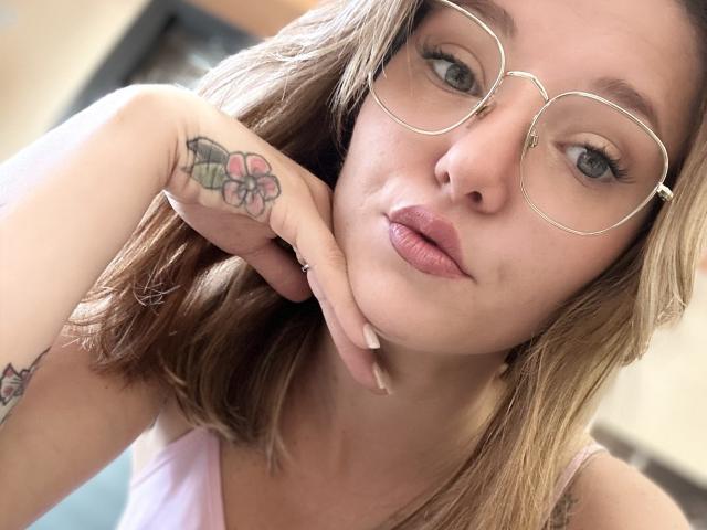 Babygirl77 with glasses, pink lipstick, blonde hair, tattoos on hand and forearm, pink tank top, looking at camera, hand under chin, light skin, soft expression, subtle makeup.