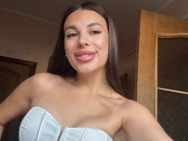 BlackFatal met lang donker haar, lichtroze lippenstift en een wit topje zonder bandjes, waarbij kleine borsten en een schattige glimlach zichtbaar zijn, staand voor een houten deur..