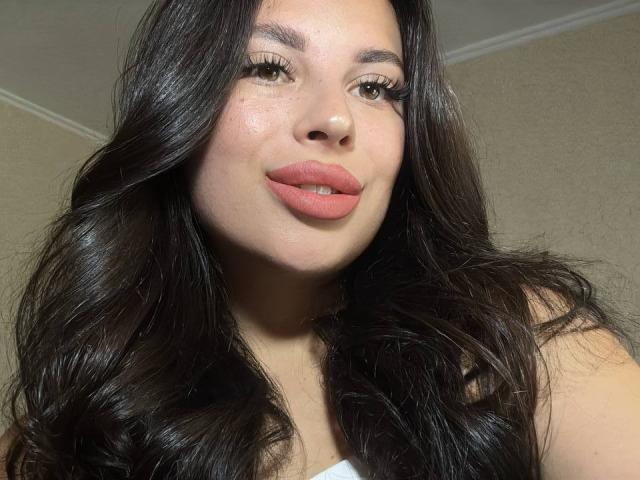 BlackFatal, con cabello negro largo y ondulado, labios rosados y ojos marrones, mirando hacia abajo a la cámara con una ligera sonrisa. Tiene un tono de piel claro y pecas visibles..