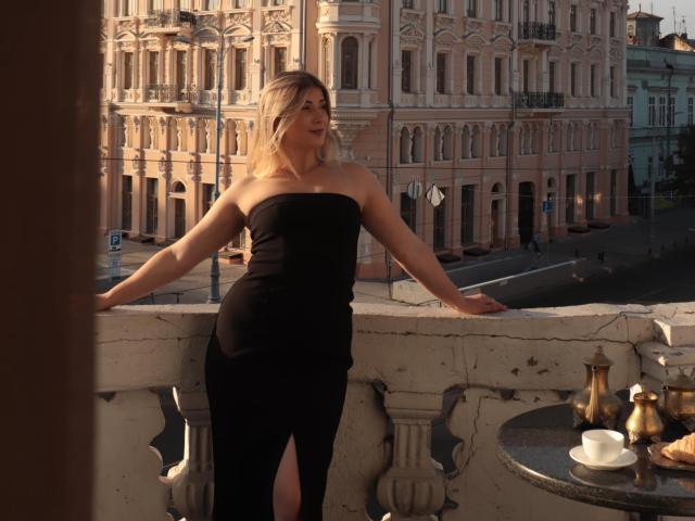 Cassandra Good, blond, trägt ein schwarzes, trägerloses Kleid mit einem Ausschnitt an den Oberschenkeln und steht auf einem Balkon, die Arme auf der Brüstung abgestützt. Es ist Abendsonne, und es steht ein kleiner, runder Tisch mit einem Tee-Service da..