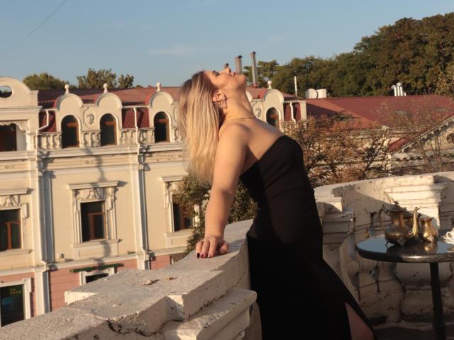 Cassandra Good lehnt sich auf einem Balkon zurück, ihr blondes Haar weht im Wind, sie trägt ein schwarzes Kleid ohne Träger, ihre Brüste sind leicht sichtbar, ihr Mund steht in einem Stöhnen offen, Sonnenlicht liegt auf ihrer Haut..