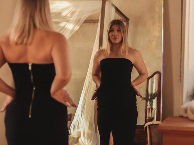 Cassandra, met blond haar, draagt een zwarte jurk zonder bandjes en staat voor een spiegel. Ze toont haar rug en haar reflectie, met haar handen op haar heupen..