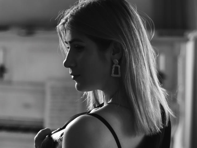 Schwarz-weißes Foto von CassandraGood, im Profil, mit blonden Haaren, die ein trägerloses Kleid trägt, einen Träger in der Hand hält, mit Diamant-Ohrringen, weiches Licht, leicht geöffneten Lippen, dezenter Make-up, elegant, sexy..
