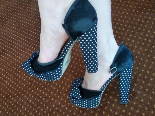 Los pies de Nurri, calzados con tacones altos negros con lunares, de tacón grueso y parte superior de terciopelo, muestran una piel pálida y una pulsera de plata en el tobillo, y están sobre un suelo cubierto de alfombra marrón..
