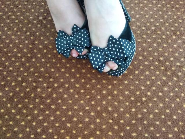 Nurri's pale feet in black, white polka-dotted, bow-adorned shoes, showing pink toenails, standing on brown, yellow-patterned carpet.