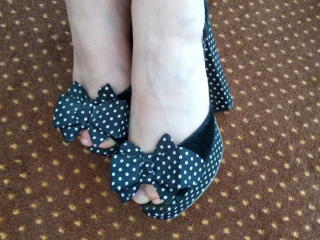 Nurris Füße in schwarzen, gepunkteten High Heels mit großen Schleifen, die blasse Haut und leicht sichtbare Zehen zeigen, stehen auf einem braunen, gemusterten Teppich..